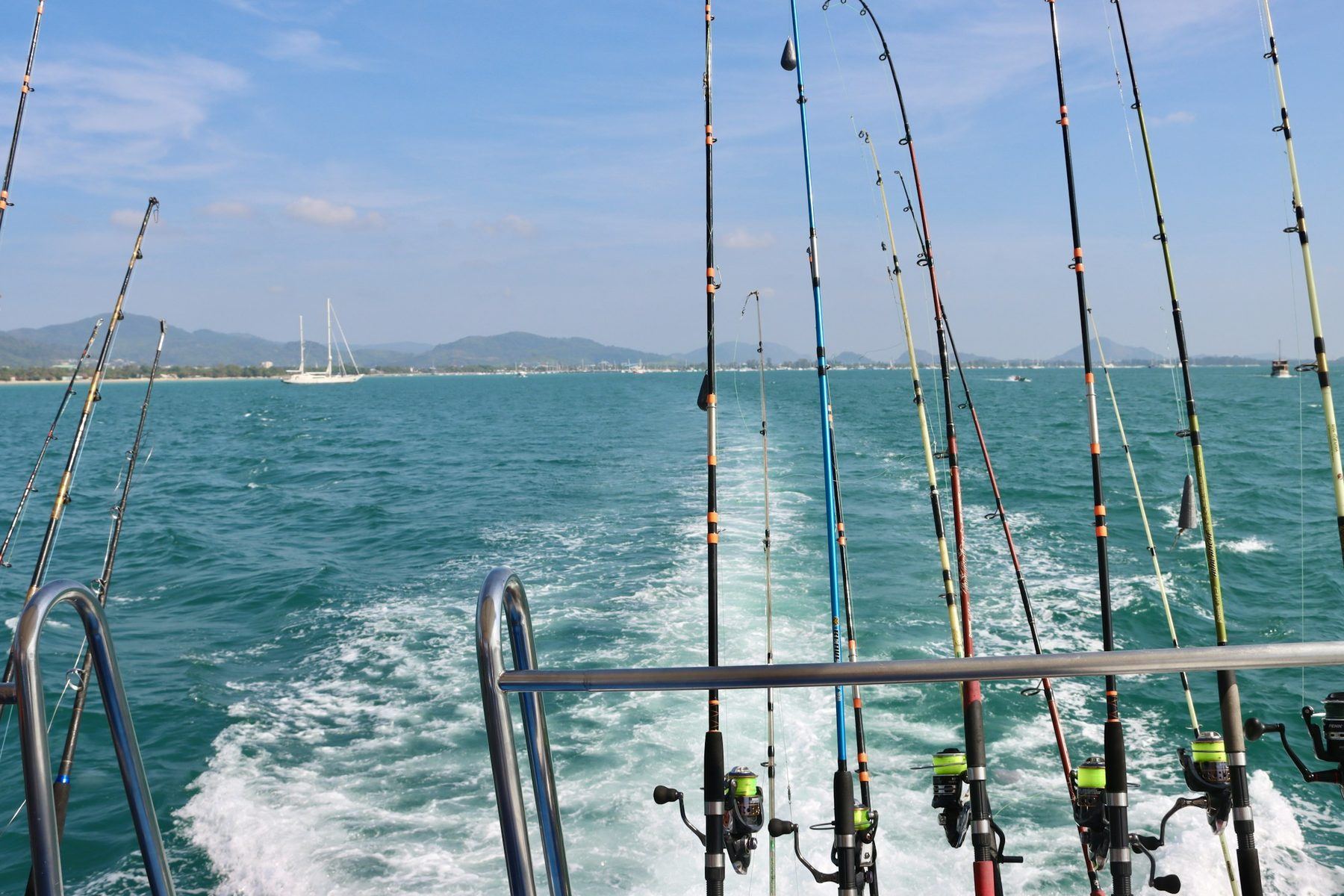 A row of trolling rods along a boat's stern rail over turquoise water and white wake